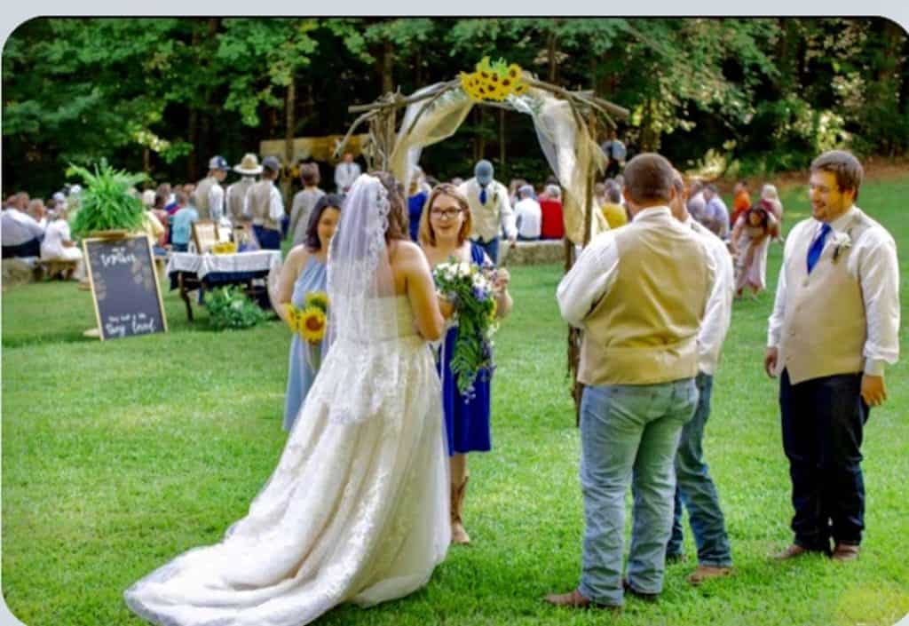 Wedding ceremony outdoors with guests, bride, and groom under a decorated arch in a lush green setti.